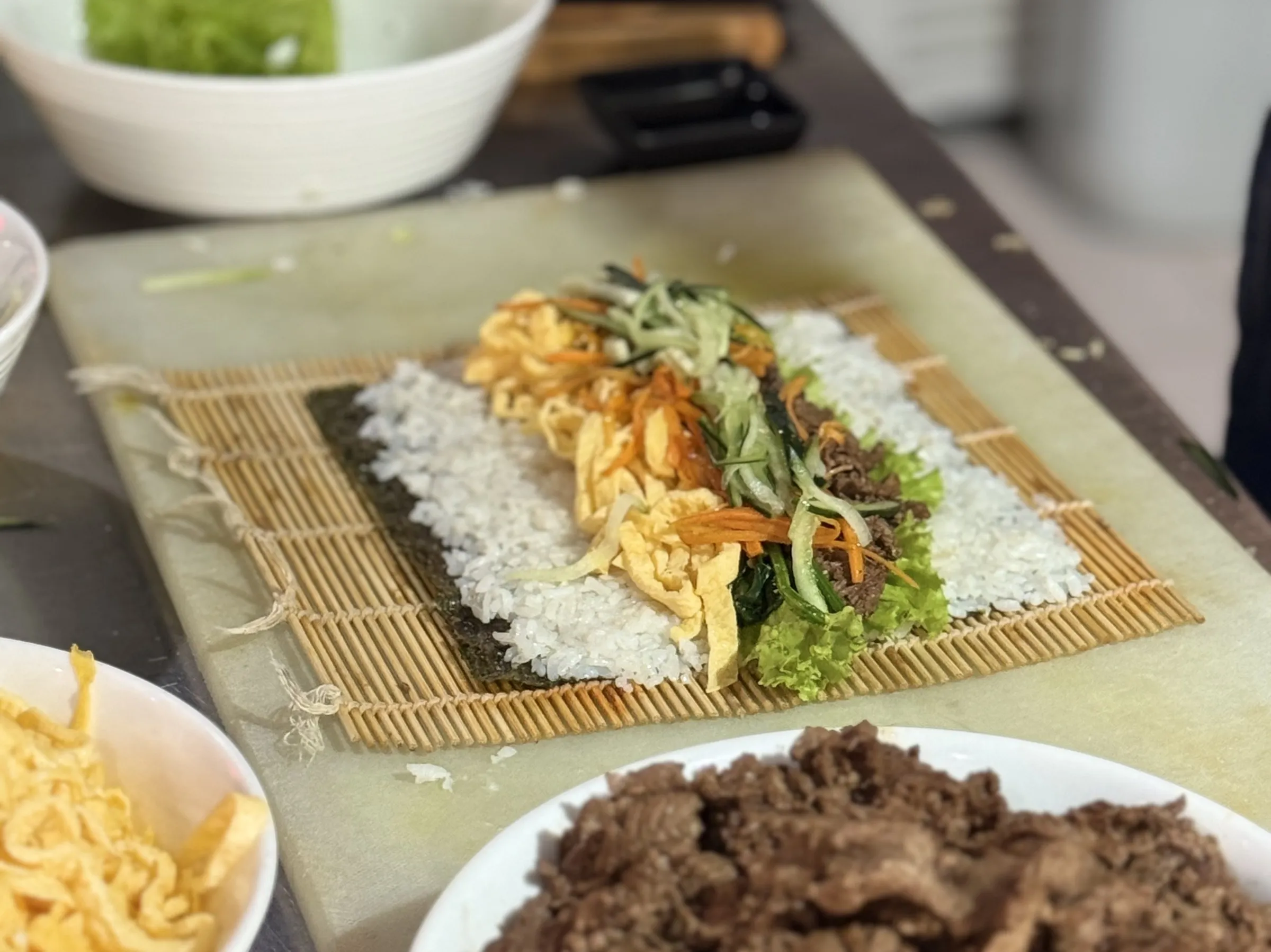 Kimbap a medio armar sobre una esterilla de bambú: arroz, huevo, zanahoria, pepino y carne.