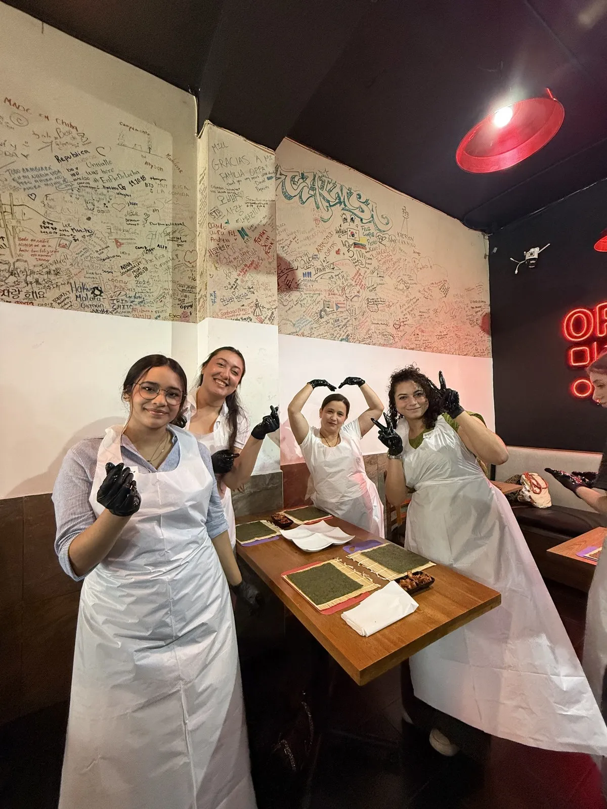 Cuatro participantes con delantal junto a una mesa con esterillas y alga para kimbap; una hace un corazón con los brazos.